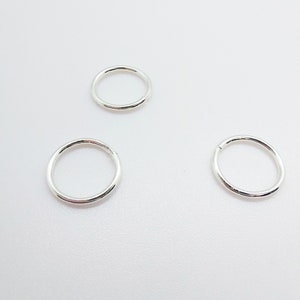 Puede incluir: Tres peque&ntilde;os pendientes de aro plateados se muestran sobre un fondo blanco. Los pendientes son circulares, con un acabado liso y pulido. Los aros est&aacute;n espaciados uniformemente, mostrando su dise&ntilde;o simple y minimalista.