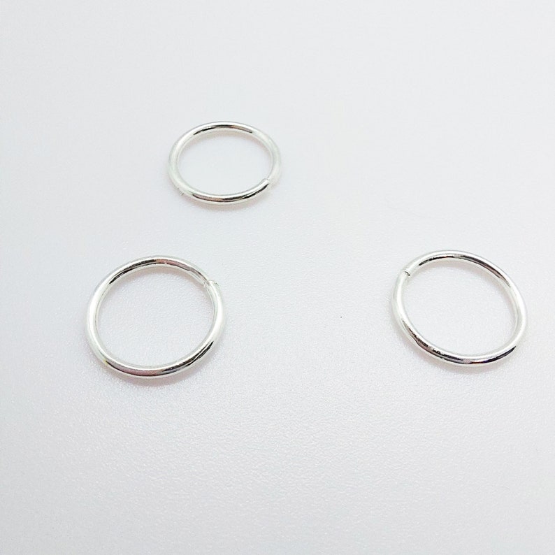 Puede incluir: Tres peque&ntilde;os pendientes de aro plateados se muestran sobre un fondo blanco. Los pendientes son circulares, con un acabado liso y pulido. Los aros est&aacute;n espaciados uniformemente, mostrando su dise&ntilde;o simple y minimalista.
