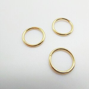 Puede incluir: Tres pendientes de aro circulares de color dorado sobre una superficie blanca. Los pendientes son sencillos, con un acabado liso y pulido. Los aros son de tama&ntilde;o y forma constantes, lo que sugiere un dise&ntilde;o de joyer&iacute;a minimalista.