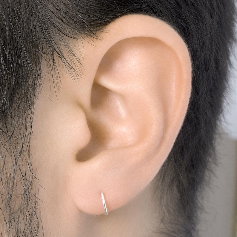 May include: A small, thin, circular silver hoop earring worn in the earlobe. The earring is a simple, polished metal ring with a seamless design, providing a minimalist and understated look that sits flush against the skin of the earlobe.