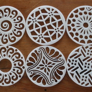 Op de afbeelding: Zes witte ronde sjablonen met verschillende geometrische en bloemmotieven. De sjablonen zijn gemaakt van een dun, flexibel materiaal en zijn perfect voor het creëren van unieke patronen op verschillende oppervlakken.
