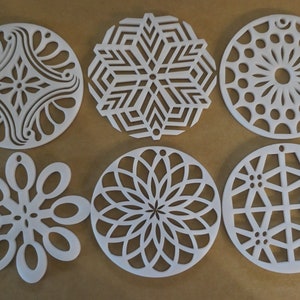 Op de afbeelding: Zes witte, lasergesneden, ronde ornamenten met ingewikkelde geometrische patronen. De patronen omvatten een sneeuwvlok, een bloem, een zonnestralen en een geometrisch patroon.