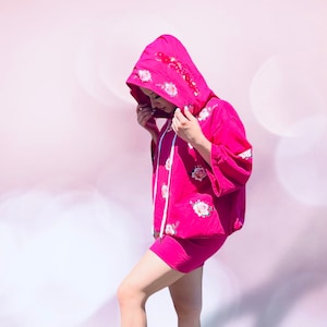 Puede incluir: Una sudadera con capucha rosa con estampado floral y una falda rosa a juego. La modelo lleva zapatillas blancas con cadenas doradas.