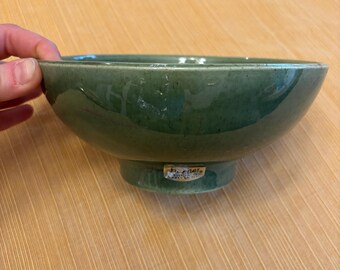 May include: A green ceramic bowl with a wide rim and a small pedestal base. The bowl has a matte finish and a subtle speckled texture. The bottom of the bowl has a label that reads "H-one's, Made in Japan".