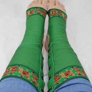 May include: Green leather leg wraps with decorative patterned trim featuring red and gold figures. The wraps are laced up the side and worn over blue jeans. The image shows the wraps from the ankles to the knees.