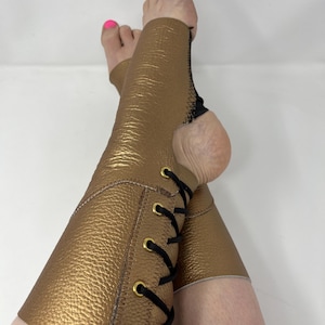 May include: Gold-coloured leg wraps with a textured finish and black lacing. The wraps have a cutout for the heel and are stitched with black thread. The lacing is secured with gold-coloured eyelets.