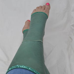 May include: Teal leg warmers with a glitter trim, worn over blue jeans. The person's feet are visible, with pink nail polish on the toes. The leg warmers are made of a soft material. The glitter trim adds a touch of sparkle.