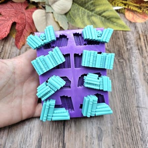 Op de afbeelding: Paarse siliconen mal met acht holtes in de vorm van stapels boeken. De mal is gevuld met lichtblauwe hars, wat stapels boeken simuleert.