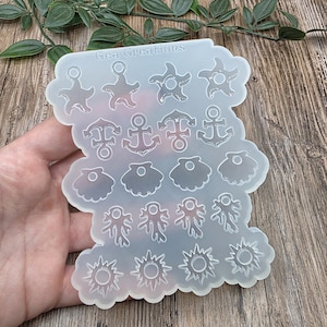 Peut inclure: Un moule en silicone transparent avec diverses formes sur le thème de la mer, notamment des étoiles de mer, des ancres, des coquillages, du corail et des soleils. Le moule porte le texte "Gea Creations" en haut. Les formes sont destinées aux projets de bricolage.