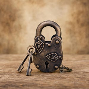 Puede incluir: Un candado de metal en tono bronce con un patrón de lágrima en relieve en su superficie frontal. Dos llaves a juego están unidas al arco, y una cadena pequeña y delicada con un diminuto dije en forma de corazón está conectada a la base.