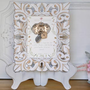 May include: An intricately carved white and gold picture frame featuring a decorative lock and two heart-shaped padlocks. The frame displays a printed card with text and floral details. The frame is supported by a stand.