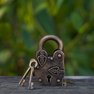 Puede incluir: Un candado de latón antiguo con intrincados diseños florales y dos llaves. El candado está sobre una superficie de madera con un fondo verde borroso.