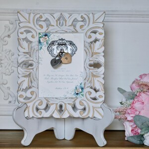 May include: A white, intricately carved picture frame with gold accents, displaying a card with floral details and two heart-shaped lock charms. The frame is supported by a white easel, creating a vintage feel.