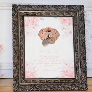 May include: Framed print with a copper-toned lock and heart charms. The print has a floral design and a quote from Matthew 19:6. The dark brown frame has detailed carvings. The print is suitable for a wedding or anniversary.