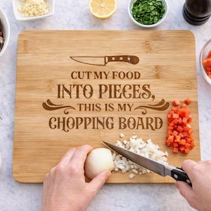 Könnte beinhalten: Ein hölzernes Schneidebrett mit dem Text "CUT MY FOOD INTO PIECES, THIS IS MY CHOPPING BOARD". Zutaten wie Pilze, Tomaten und Kräuter umgeben das Brett. Ein Messer wird zum Zerkleinern einer Zwiebel verwendet.