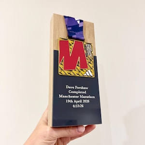 Puede incluir: Placa de madera con una medalla del Maratón de Manchester. La medalla dorada tiene una "M" roja y una cinta morada. La placa tiene una sección negra con el texto "Dave Forshaw Completed Manchester Marathon 19th April 2026 4:53:26".