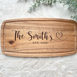 May include: Wooden rectangular chopping board with a carved groove around the edge. The text "The Smith's" is engraved in a cursive font, with a heart symbol and "EST. 1989" below. The board has a natural wood grain.