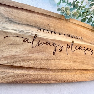 Op de afbeelding: Houten snijplank met gegraveerde rand en de tekst "MEATS & CHEESES always pleases". De plank is gemaakt van lichtbruin hout en is geschikt voor het serveren van hapjes, ongeveer 30 cm lang.