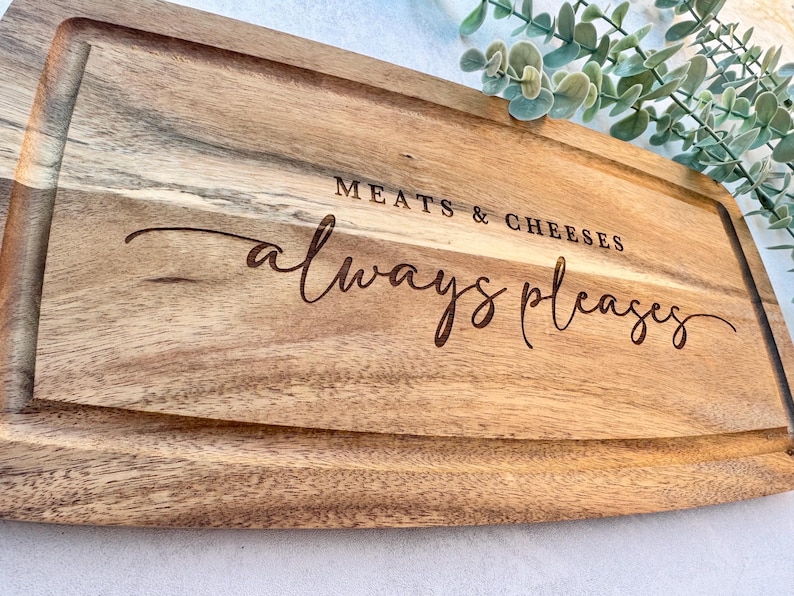 Op de afbeelding: Houten snijplank met gegraveerde rand en de tekst "MEATS & CHEESES always pleases". De plank is gemaakt van lichtbruin hout en is geschikt voor het serveren van hapjes, ongeveer 30 cm lang.