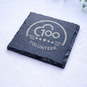 Op de afbeelding: Vierkante, donkergrijze leistenen onderzetter met de tekst "100 VOLUNTEER" in lichtgrijs. Het ontwerp bevat een wolkvorm, vijf sterren en horizontale lijnen. De onderzetter ligt op een witte ondergrond.
