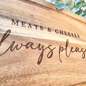 Op de afbeelding: Houten snijplank met de gegraveerde tekst "MEATS & CHEESES always pleases". De plank heeft een rechthoekige vorm met een groef rond de rand. De houtnerf is zichtbaar en de plank is lichtbruin.