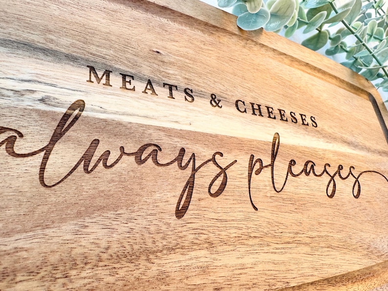 Op de afbeelding: Houten snijplank met de gegraveerde tekst "MEATS & CHEESES always pleases". De plank heeft een rechthoekige vorm met een groef rond de rand. De houtnerf is zichtbaar en de plank is lichtbruin.