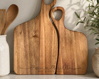 May include: Two light brown wooden serving boards with handles, featuring a natural wood grain pattern. The boards are engraved with the names Ross and Rachel, along with the date 22nd August 2028, written in a cursive script font.