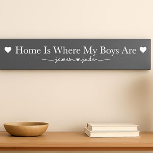 May include: A dark grey wooden sign with the text "Home Is Where My Boys Are" in white, with two white hearts. The names "james & jude" are written in cursive. The sign is displayed above a wooden table.