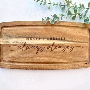 Op de afbeelding: Rechthoekige houten serveerplank met een sapgroef en de gegraveerde tekst "MEATS & CHEESES always pleases". De plank is gemaakt van lichtbruin hout en is omgeven door groen.
