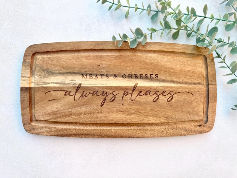 Op de afbeelding: Rechthoekige houten serveerplank met een sapgroef en de gegraveerde tekst "MEATS & CHEESES always pleases". De plank is gemaakt van lichtbruin hout en is omgeven door groen.