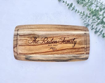 Puede incluir: Tabla de cortar rectangular de madera con borde tallado. El texto "The Roden Family EST 2007" está grabado en una fuente decorativa. Un pequeño diseño de hoja está debajo del texto. La tabla es de madera marrón clara y oscura.
