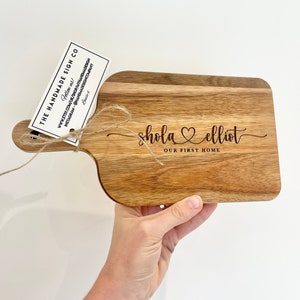 Può includere: Un tagliere in legno con manico, inciso con i nomi "shola elliot" e il testo "OUR FIRST HOME".