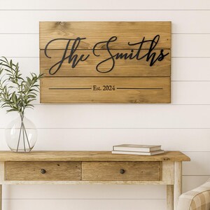 May include: A wooden sign with the name "The Smiths" in a cursive font, and the text "Est. 2024" below. The sign is mounted on a white wall above a wooden console table. A clear glass vase with greenery sits on the table.