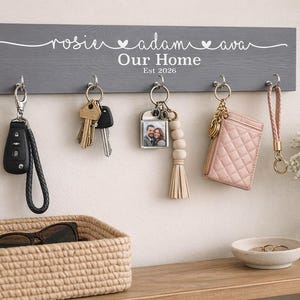 May include: A grey wooden key holder with the names "rosie adam ava" and "Our Home Est 2026" in white script. It has six silver hooks holding keys, a car key fob, a pink wallet, and a wristlet. A woven basket and a small bowl are on a wooden shelf.