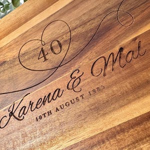 Puede incluir: Tabla de cortar de madera con texto grabado y forma de corazón. El texto dice "40 Karena & Mal 10th August 1985". La tabla tiene un patrón de vetas de madera natural.