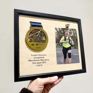 Könnte beinhalten: Ein gerahmtes Display mit einer goldenen Manchester Marathon 2023 Medaille und einem Foto einer Läuferin. Die Medaille hat ein blaues Band. Das Foto zeigt eine Läuferin in einem gelben Shirt. Text auf dem Rahmen enthält den Namen der Läuferin, Renndetails und Zeit.
