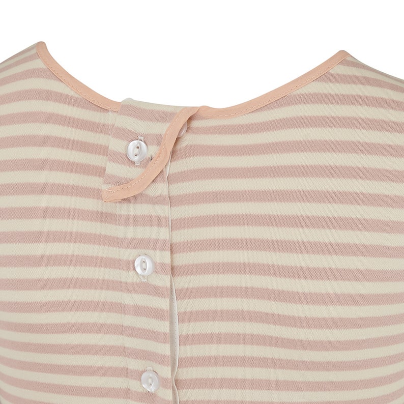 May include: Close-up of a striped garment in cream and pale pink. The fabric features horizontal stripes and a row of white buttons down the front. The neckline and button placket are trimmed with a soft peach-colored fabric.