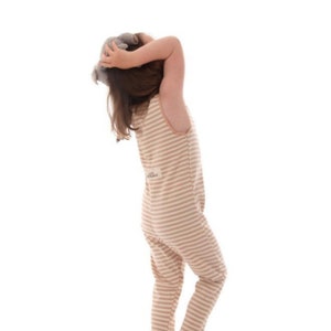 May include: A beige and white striped jumpsuit with a round neckline. The jumpsuit has a small patch on the back and is worn by a person holding a stuffed animal. The background is white.