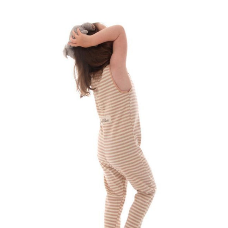 May include: A beige and white striped jumpsuit with a round neckline. The jumpsuit has a small patch on the back and is worn by a person holding a stuffed animal. The background is white.