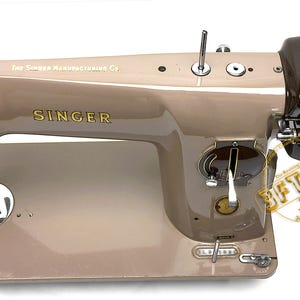 May include: A vintage Singer sewing machine in a beige and brown color scheme. The machine features the Singer logo in gold lettering and the text "The Singer Manufacturing Co." The machine has various dials and levers.