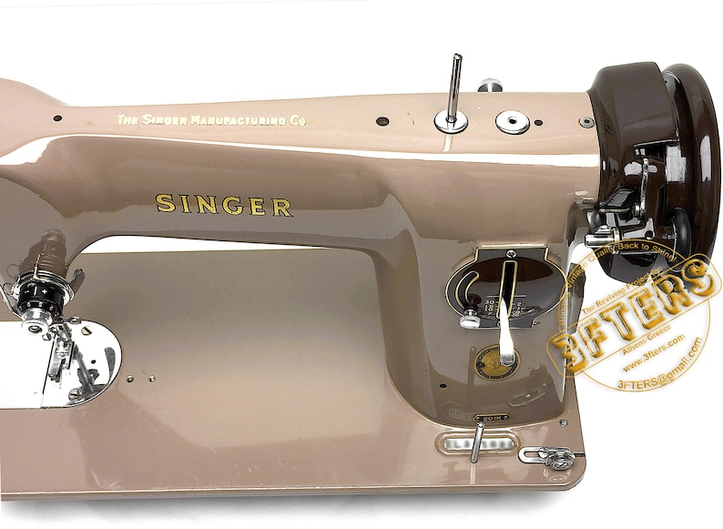 May include: A vintage Singer sewing machine in a beige and brown color scheme. The machine features the Singer logo in gold lettering and the text "The Singer Manufacturing Co." The machine has various dials and levers.