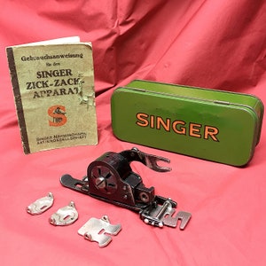 Könnte beinhalten: Eine grüne Metallbox mit dem Wort "SINGER" darauf gedruckt. Ein schwarzer Nähmaschinenaufsatz mit einem Metallfuß und eine kleine Gebrauchsanweisung mit dem Titel "Gebrauchsanweisung für den SINGER ZICK-ZACK APPARAT" sind ebenfalls abgebildet.