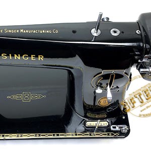 Puede incluir: M&aacute;quina de coser Singer negra con letras y detalles dorados. Los textos "THE SINGER MANUFACTURING CO." y "SINGER" son visibles. La m&aacute;quina tiene un acabado brillante y adornos dorados decorativos.