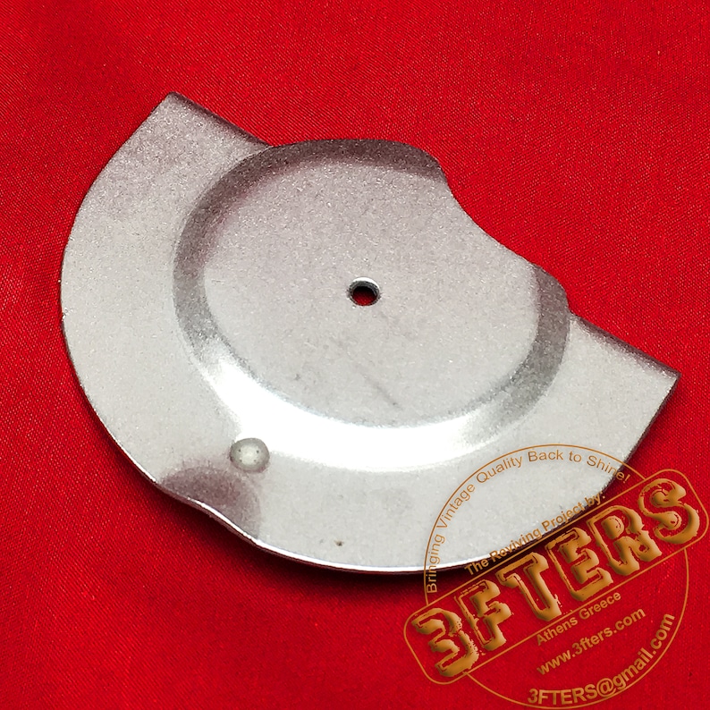 May include: A silver metal disc with a hole in the center and a smaller hole on the side. The disc is on a red surface. The text on the disc reads: 'Bringing Vintage Quality Back to Shirel The Reviving Project by: 3FTERS Athens Greece www.3fters.com 3FTERS@gmail.com'