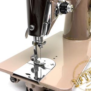 May include: A vintage sewing machine with a brown and beige color scheme. The machine features a shiny chrome needle and a beige base. The machine has a classic design, with a focus on functionality and durability. The machine is likely used for sewing and crafting.