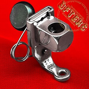 Könnte beinhalten: Ein Vintage-Metall-Nähmaschinen-Teil mit Feder und einem schwarzen zylindrischen Teil. Das Teil hat eine sechseckige Form und einen gebogenen Sockel mit der Nummer "86294" und dem Text "SIMANOO". Der Hintergrund ist rot.