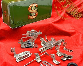 May include: A green metal box with the words "Singer" and "Naehmaschinen" printed on it. The box contains a set of vintage sewing machine attachments, including a foot pedal, a needle threader, and a variety of presser feet.