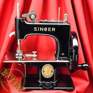Könnte beinhalten: Eine schwarze Vintage-Nähmaschine Singer mit einer goldfarbenen Namensschilder und einem schwarzen Fußpedal. Die Maschine steht auf einer roten Oberfläche.