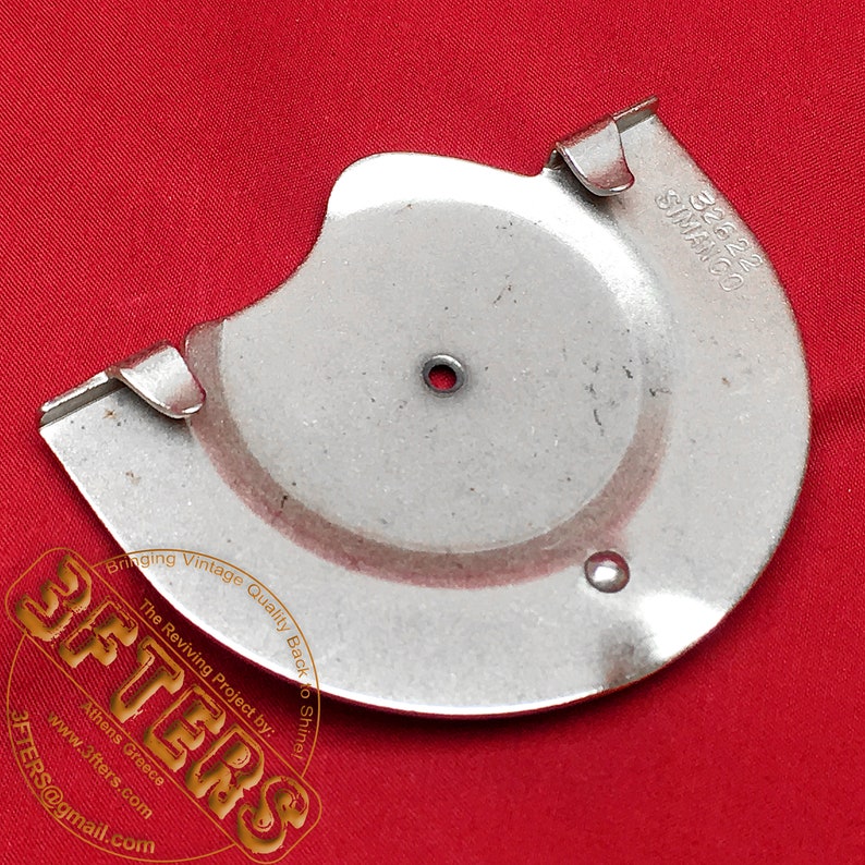 May include: A silver metal disc with a hole in the center and a curved edge. The disc has the text '32622 SIMANCO' printed on the side. The disc is on a red surface.