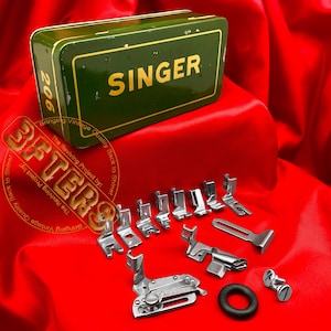 May include: A green Singer sewing machine accessory box with gold lettering and the number 206. The box is next to a collection of silver sewing machine feet and a black rubber ring, all on a red satin surface.